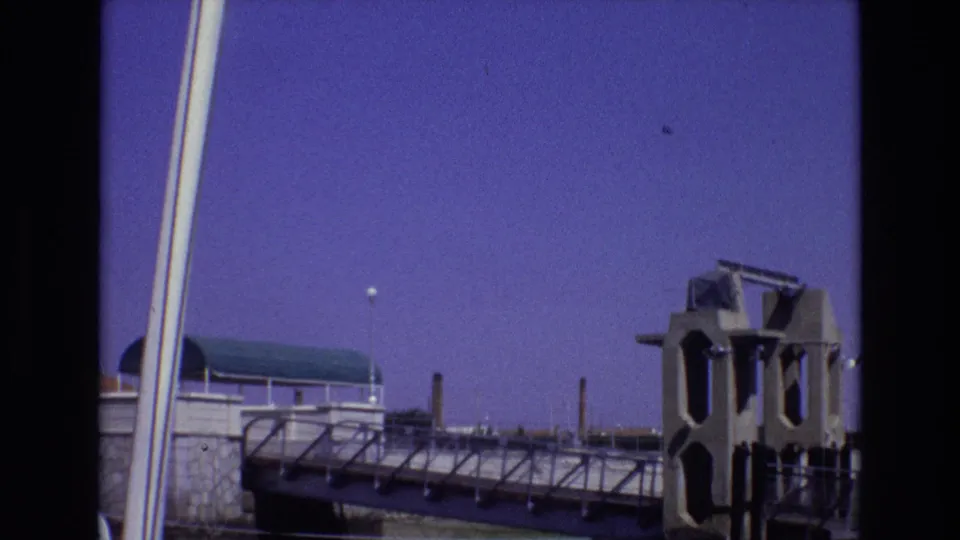 1972 Portugal 8mm frame continuing the view of a waterfront or nautical structure, with a green roof, walkway railings, smokestacks, and a concrete or metal form at right.