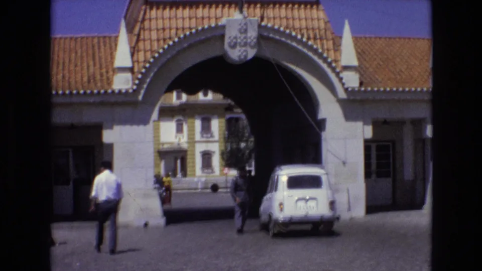 1972 Portugal 8mm frame showing a stone archway with an orange tiled roof and coat of arms, pedestrians near the entrance, and a small white car inside the gateway.