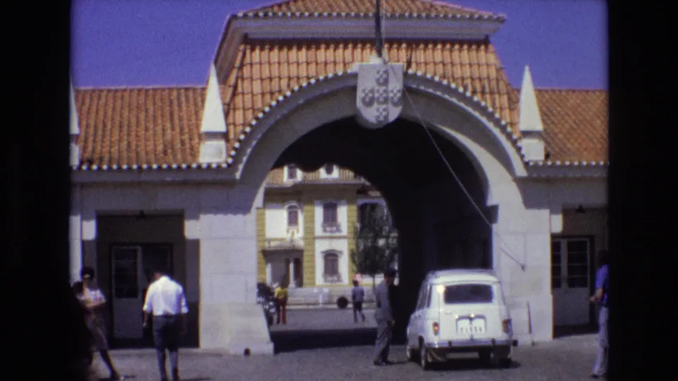1972 Portugal 8mm frame showing the ornate stone gateway from a slightly shifted angle, with pedestrians on both sides and a white compact car near the arch.