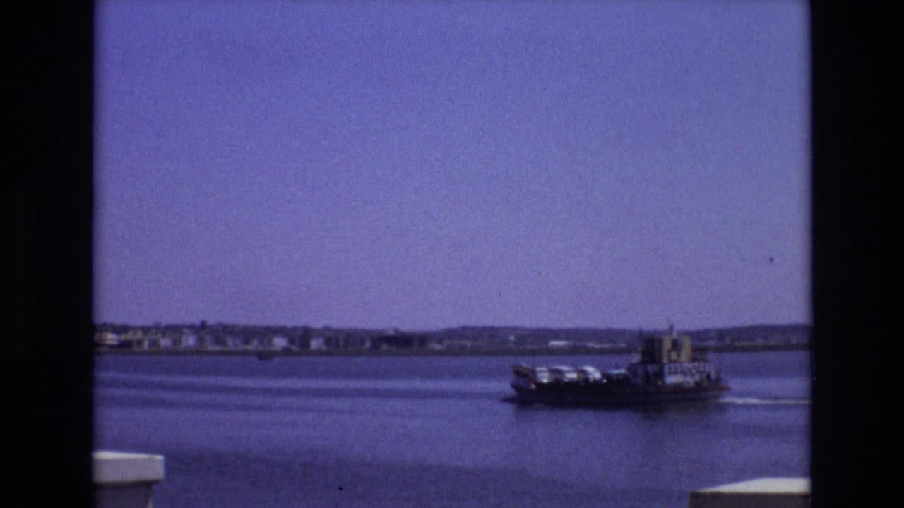 1972 Portugal 8mm frame showing a loaded ferry moving across a calm bay, with a distant shoreline, low hills, and dark film borders visible.