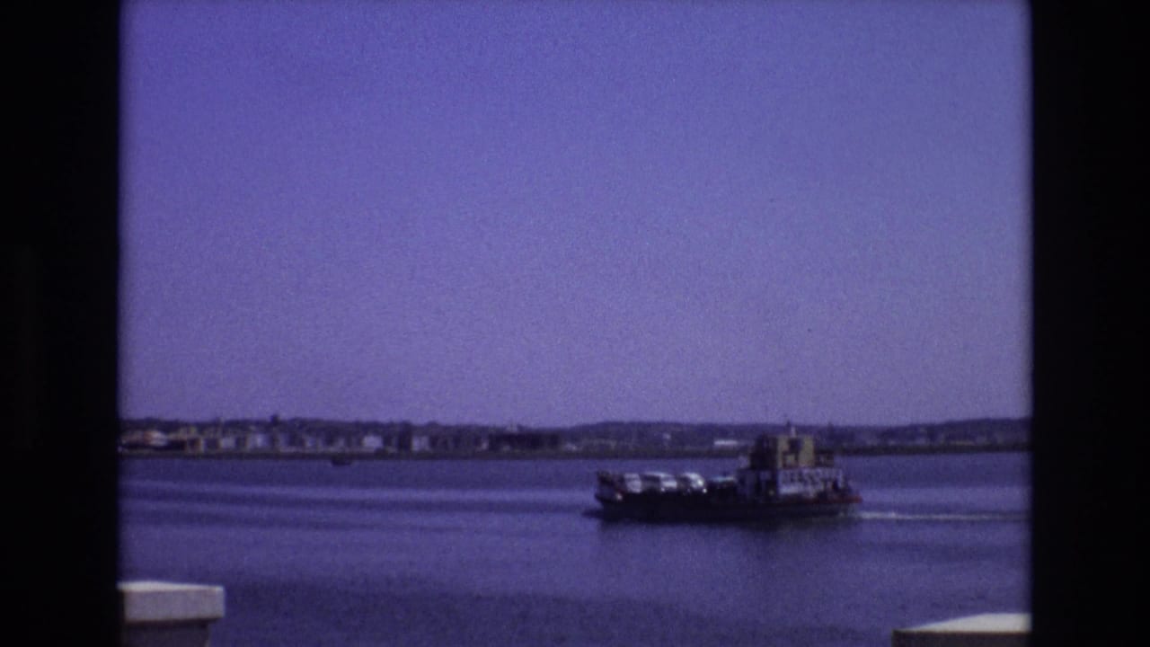 1972 Portugal 8mm frame with a ferry carrying vehicles or cargo across open bay water, framed by the scanned film edge and a distant shoreline.