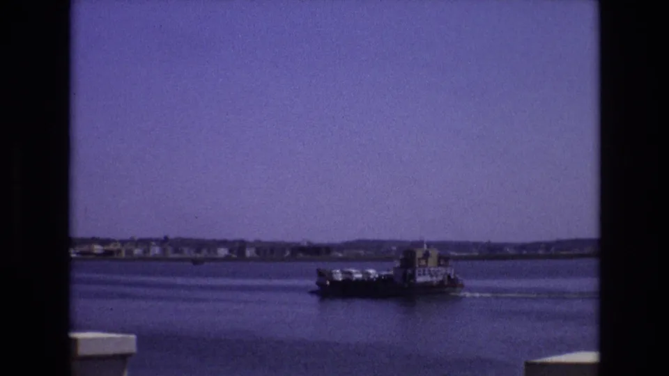 1972 Portugal 8mm frame showing the ferry farther across the bay, with rippled water, distant shoreline buildings, low hills, and a pale foreground edge.