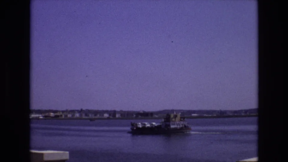 1972 Portugal 8mm frame of a loaded ferry crossing calm bay water near the edge of the frame, with distant coastal buildings and low hills beyond.