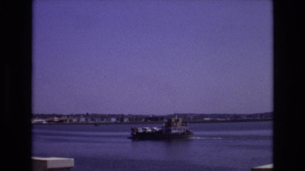 1972 Portugal 8mm frame showing a small riverboat moving across calm water, with a low shoreline and buildings in the distance.