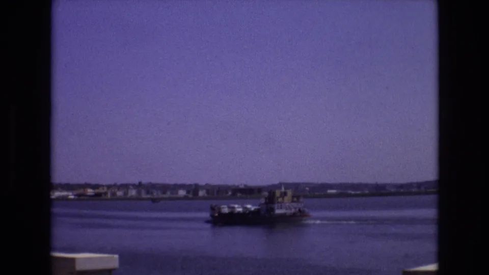 1972 Portugal 8mm frame showing a small riverboat moving across calm water, with a low shoreline and buildings in the distance.