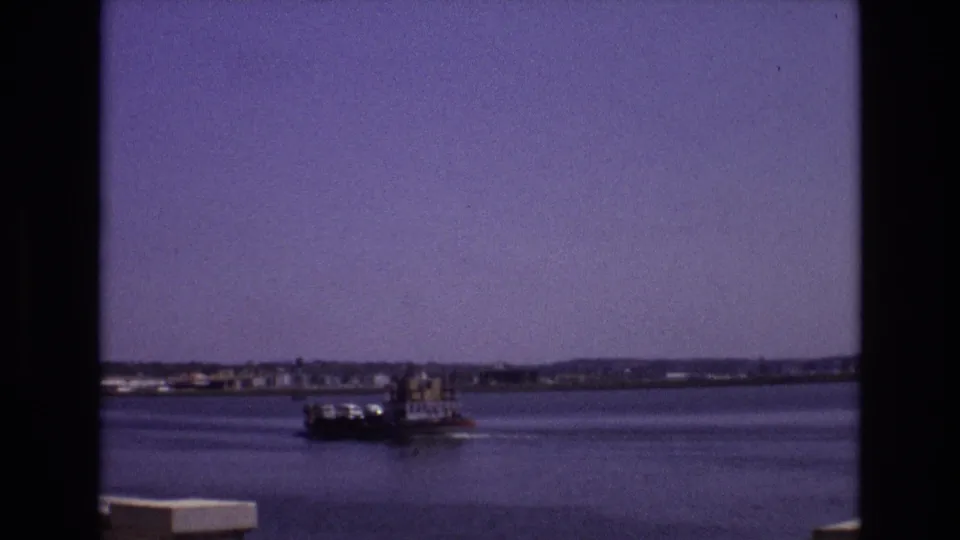1972 Portugal 8mm frame of a small boat leaving a gentle wake across a wide waterway, with a low town shoreline and hazy sky beyond.