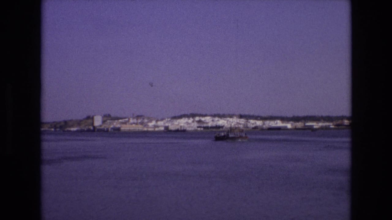 Small boat crossing calm water toward a distant shoreline town.