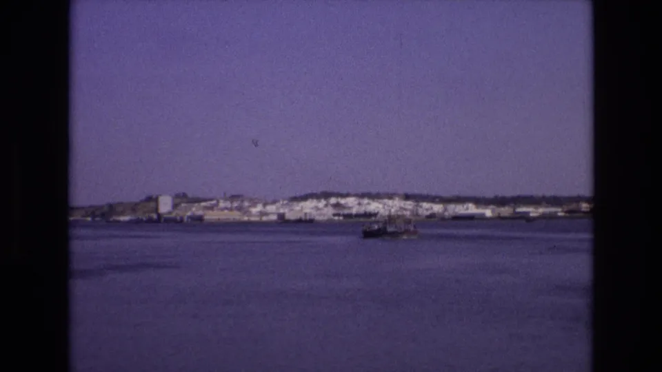 1972 Portugal 8mm frame showing a small boat on a calm bay with white buildings and low hills along the far shore.