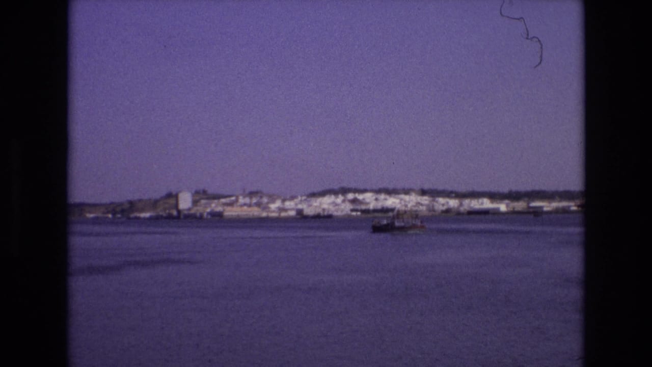 1972 Portugal 8mm frame with a small vessel moving across open water in front of a distant white coastal town.