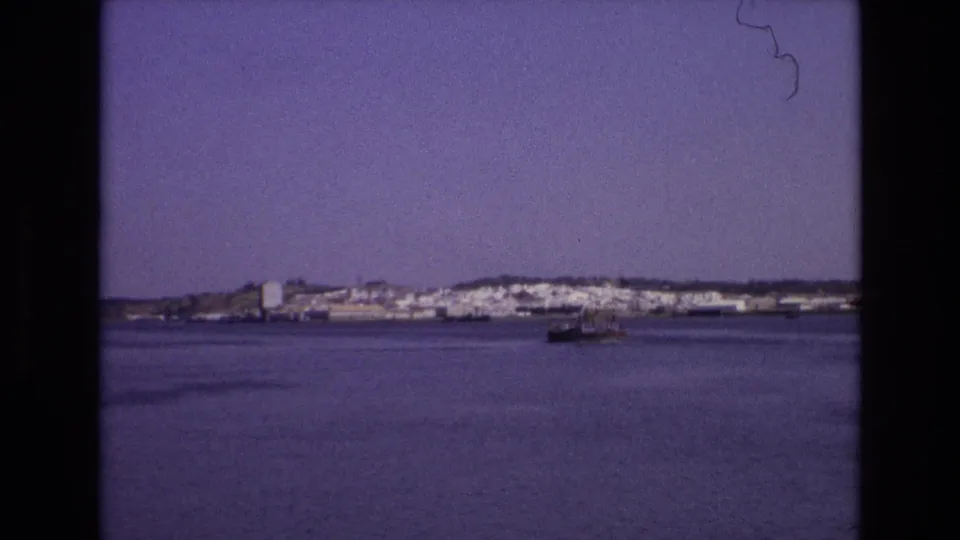 1972 Portugal 8mm frame with a small vessel moving across open water in front of a distant white coastal town.