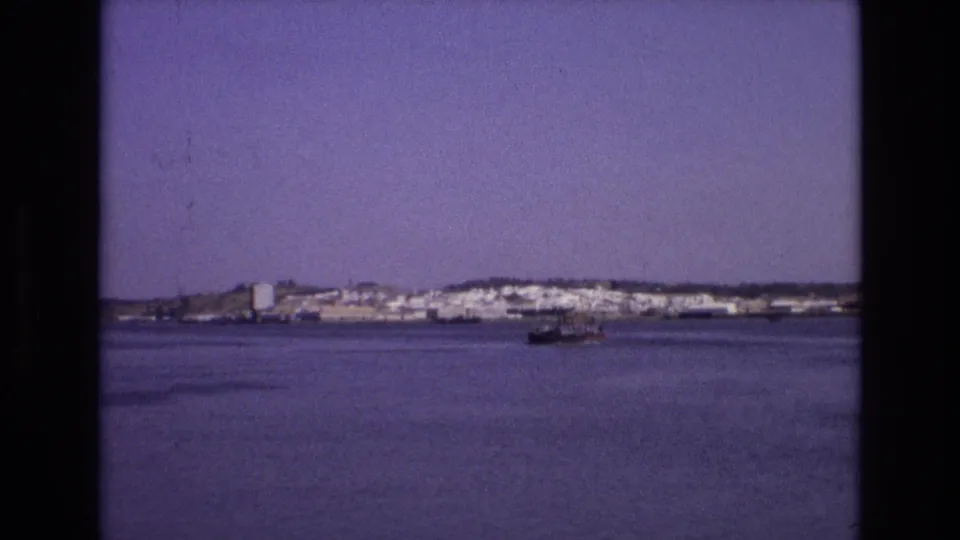1972 Portugal home-movie frame showing a boat crossing calm water below a distant shoreline of white low-rise buildings.