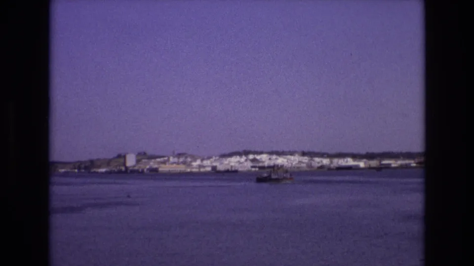1972 Portugal 8mm frame showing a small boat on a quiet bay, with a distant waterfront town and low hills under a clear sky.