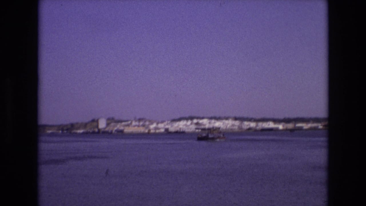 Portugal 1972 home-movie frame showing white shoreline buildings across a calm bay, with a small boat moving in the middle distance.