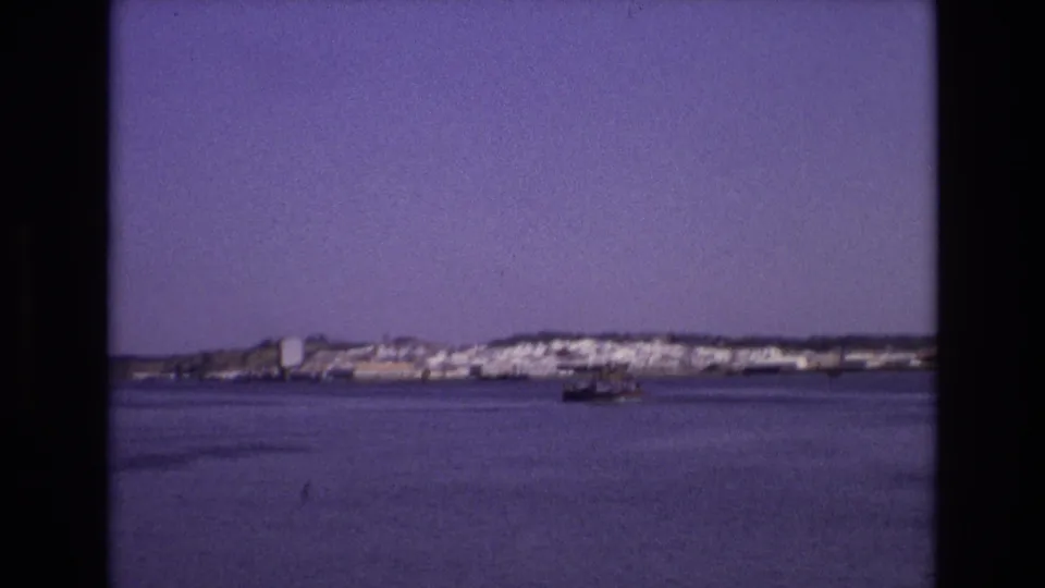 Portugal 1972 home-movie frame showing white shoreline buildings across a calm bay, with a small boat moving in the middle distance.