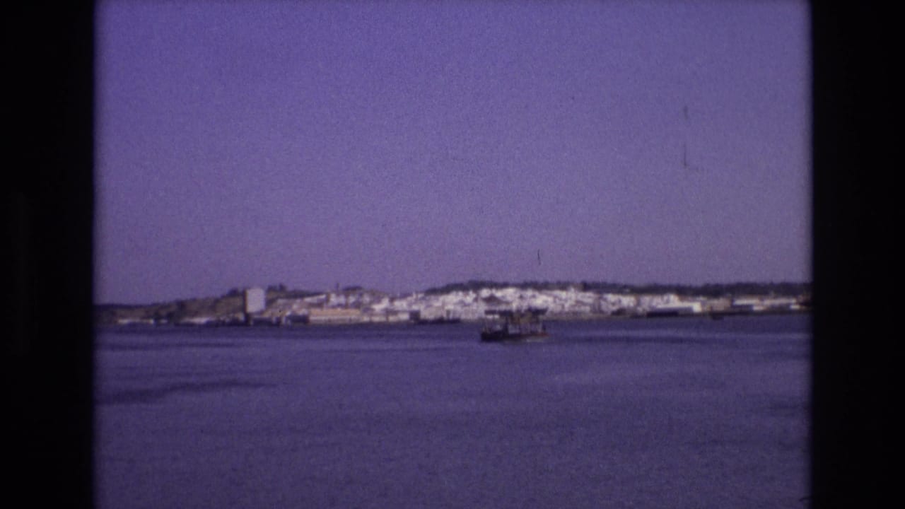 A soft 8mm view of a Portuguese coastal town across the water, with a small boat near the center of the bay under a clear sky.