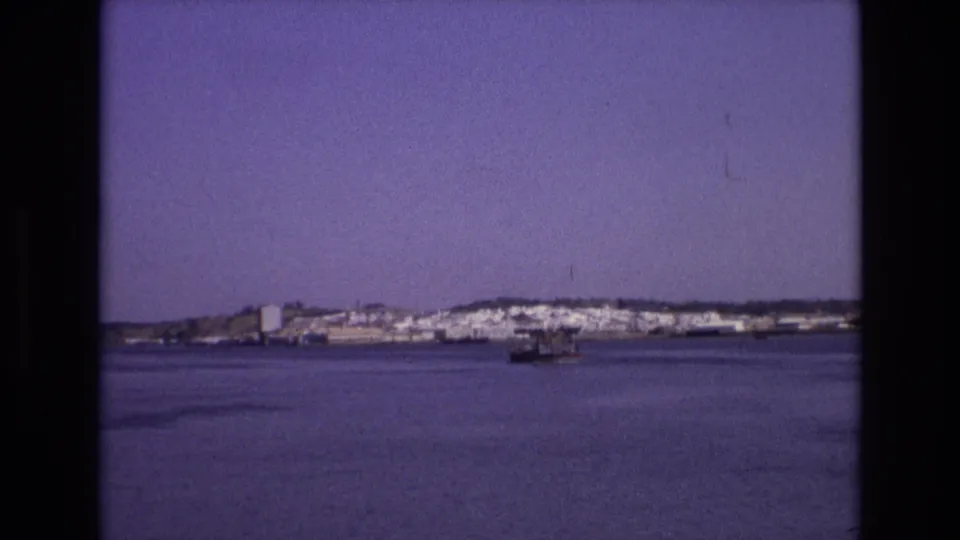 A soft 8mm view of a Portuguese coastal town across the water, with a small boat near the center of the bay under a clear sky.