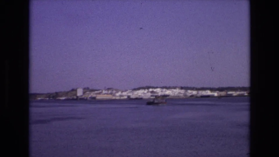 Wide 1972 Portugal travel frame of a calm bay, distant white buildings, and a small boat set against a low shoreline.
