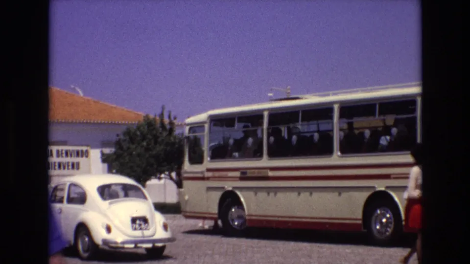 Street scene in Portugal, 1972, with a cream-and-red bus, visible passengers, a white Volkswagen Beetle, and a nearby building sign reading “Bem Vindo.”