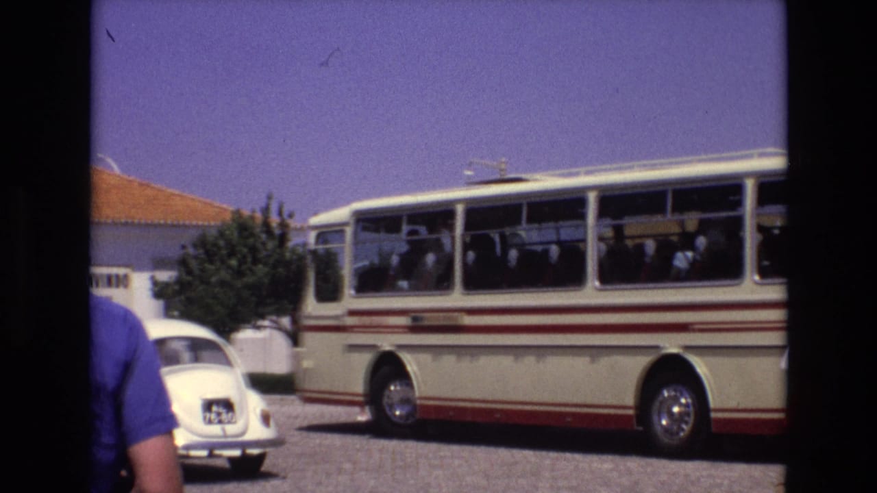1972 Portugal 8mm frame showing a cream tour bus with red stripes, seated passengers, a white Volkswagen Beetle, a terracotta-roof building, and a person partly in the foreground.