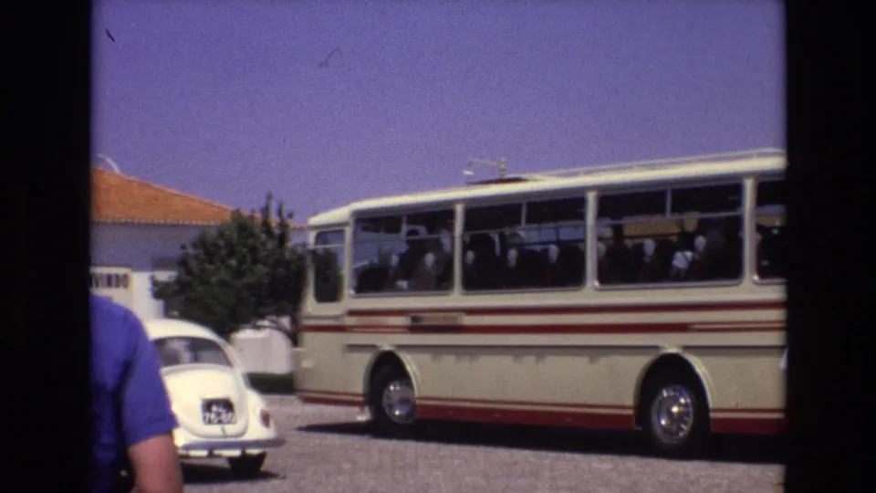 1972 Portugal 8mm frame showing a cream tour bus with red stripes, seated passengers, a white Volkswagen Beetle, a terracotta-roof building, and a person partly in the foreground.