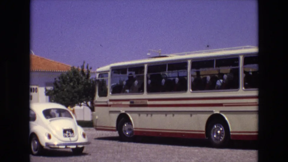 1972 Portugal 8mm frame with a white Volkswagen Beetle in the foreground, a cream tour bus with red stripes behind it, passengers in the bus, and a terracotta-roof building nearby.