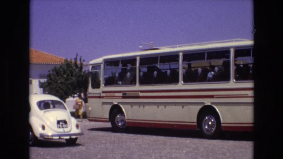 1972 Portugal home-movie frame showing the side of a cream bus with red stripes, passengers seated inside, a white Volkswagen Beetle, a tree, and a sunlit building edge.
