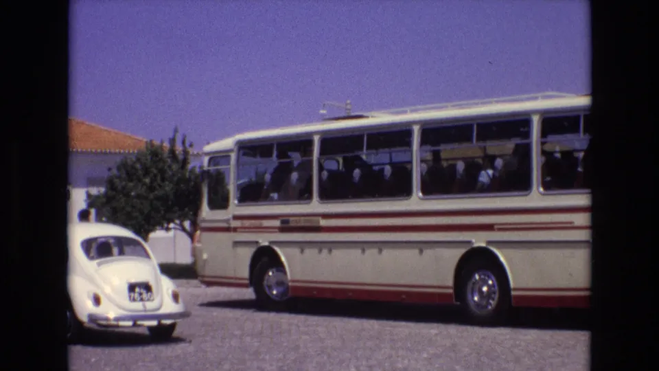 1972 Portugal home-movie frame showing a classic white-and-red tour bus with passengers visible through the windows and a white Volkswagen Beetle in the foreground.