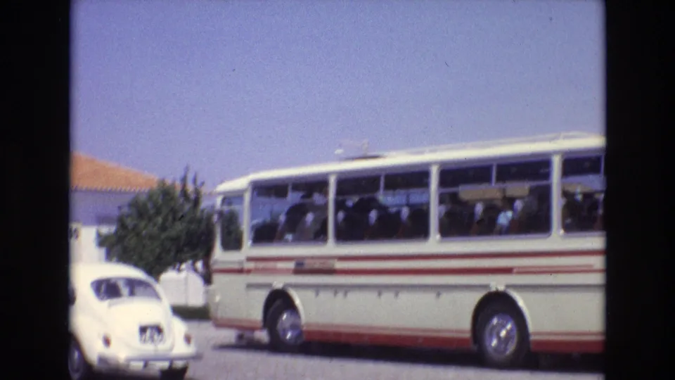 Soft 8mm frame from Portugal in 1972 showing the side of a white-and-red tour bus and the rear of a white Volkswagen Beetle at the edge of the shot.
