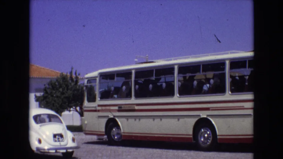 1972 Portugal home-movie frame showing a classic tour bus with visible passengers, a white Volkswagen Beetle, cobblestones, and a small tree in bright sunlight.