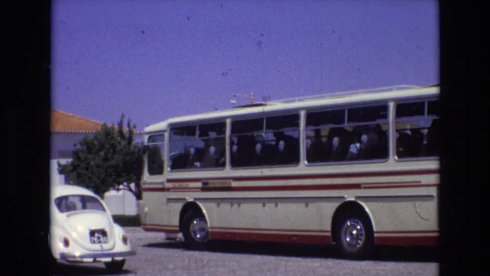 1972 Portugal travel footage frame with a white and red coach bus, visible passengers, a white Volkswagen Beetle, and sunlit roadside buildings.