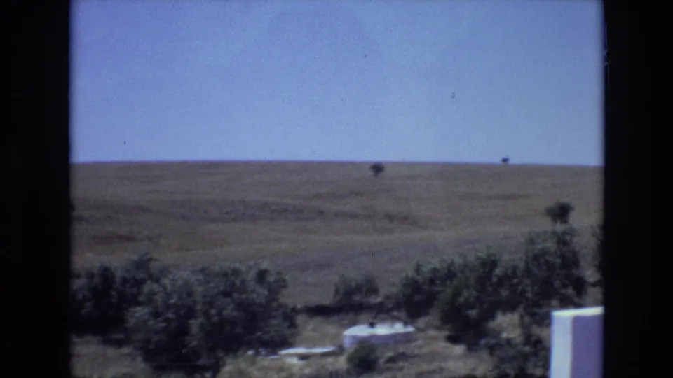1972 Portugal rural landscape frame with shrubs, scattered trees, small pale structures, and a wide open hillside under blue sky.