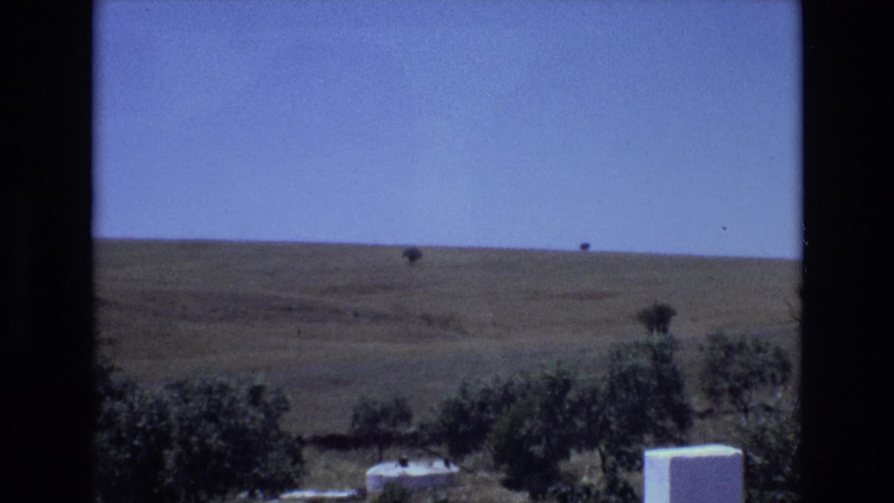 1972 Portugal countryside frame with a broad dry hill, sparse small trees on the horizon, dark foreground vegetation, and a white structure near the lower right.