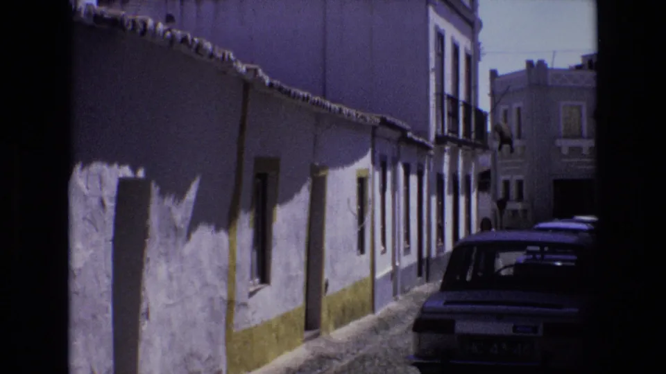 1972 Portugal street view showing whitewashed buildings with yellow lower trim, a narrow cobblestone lane, and a parked dark car with its license plate visible.