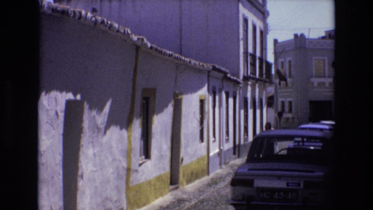 Portugal 1972 home-movie frame showing a narrow cobblestone street lined with white-washed buildings with yellow trim and a parked period car.
