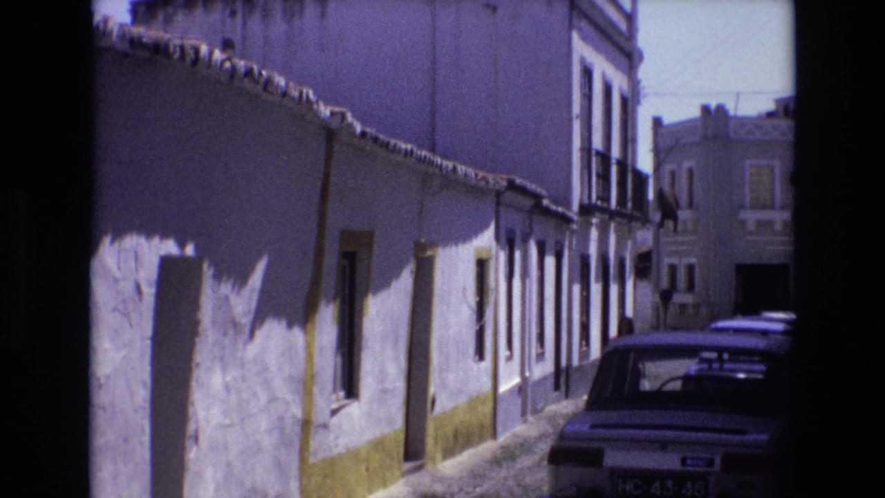Sunlit white-washed street with yellow trim and the rear of a parked car.