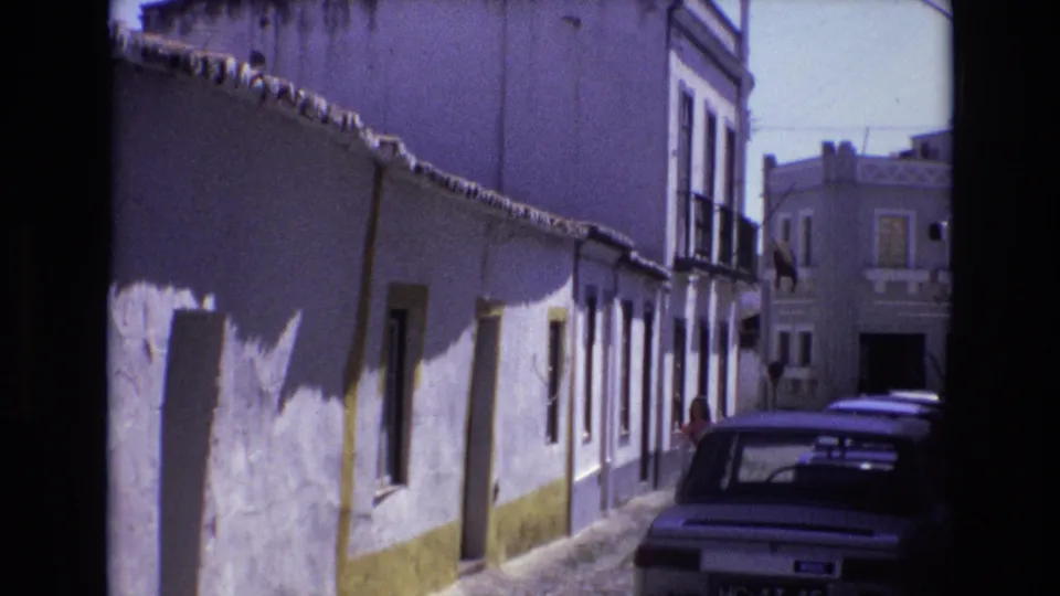Portugal 1972 8mm frame showing a white-washed cobblestone street, yellow wall trim, parked car, and a distant person near the buildings.