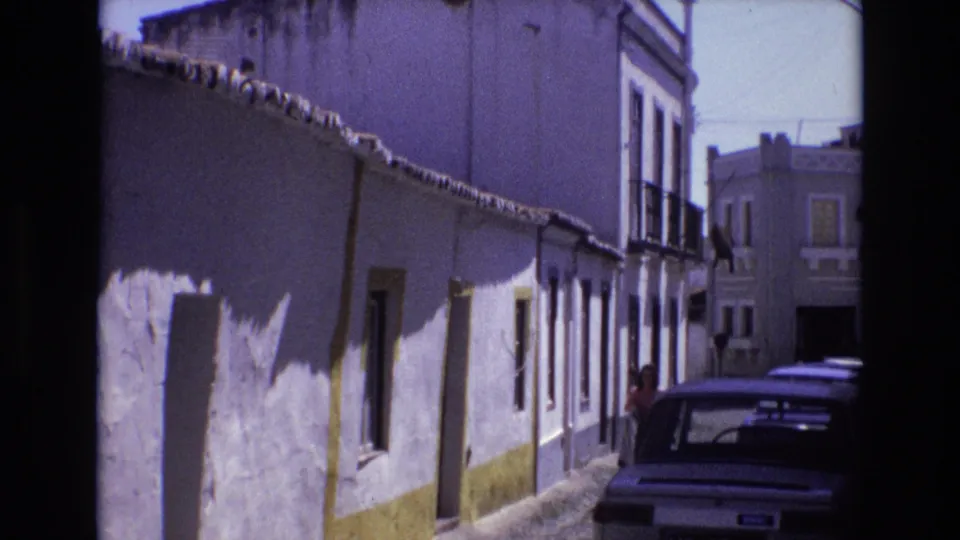 Portugal 1972 frame of a narrow cobblestone street with white-washed buildings, yellow trim, a parked car, and a person partly visible near the car.