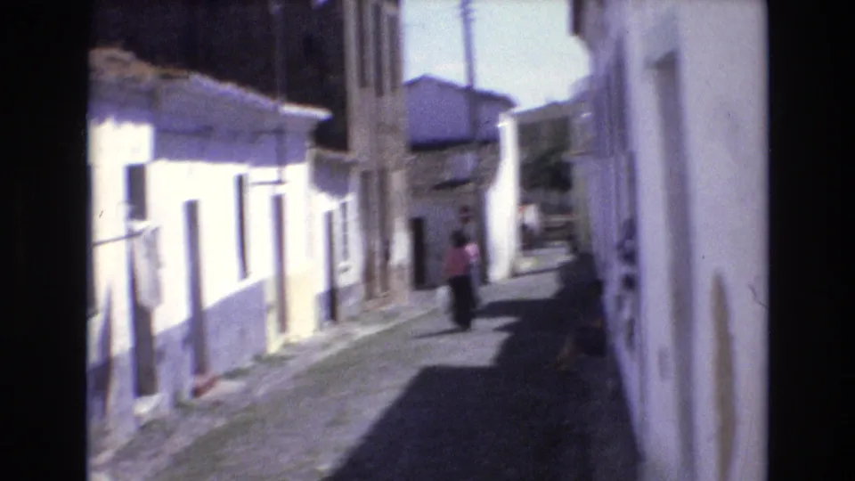 Portugal 1972 home-movie frame showing a woman in a pink top walking away along a narrow cobblestone alley lined with whitewashed buildings and strong sunlit shadows.