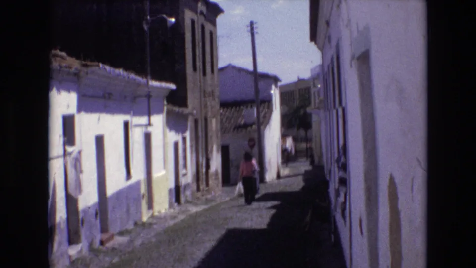 Portugal 1972 home-movie frame showing a woman carrying a bag as she walks away through a narrow cobblestone alley bordered by whitewashed walls, yellow trim, and utility poles.