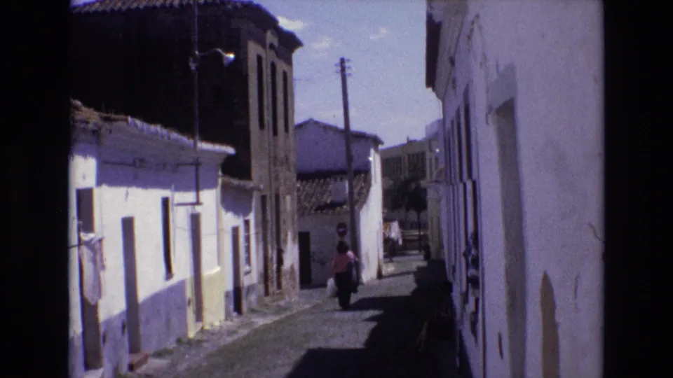Portugal 1972 home-movie frame showing the woman farther down the alley, framed by whitewashed buildings, tiled roofs, a utility pole, and a small stop sign in the distance.
