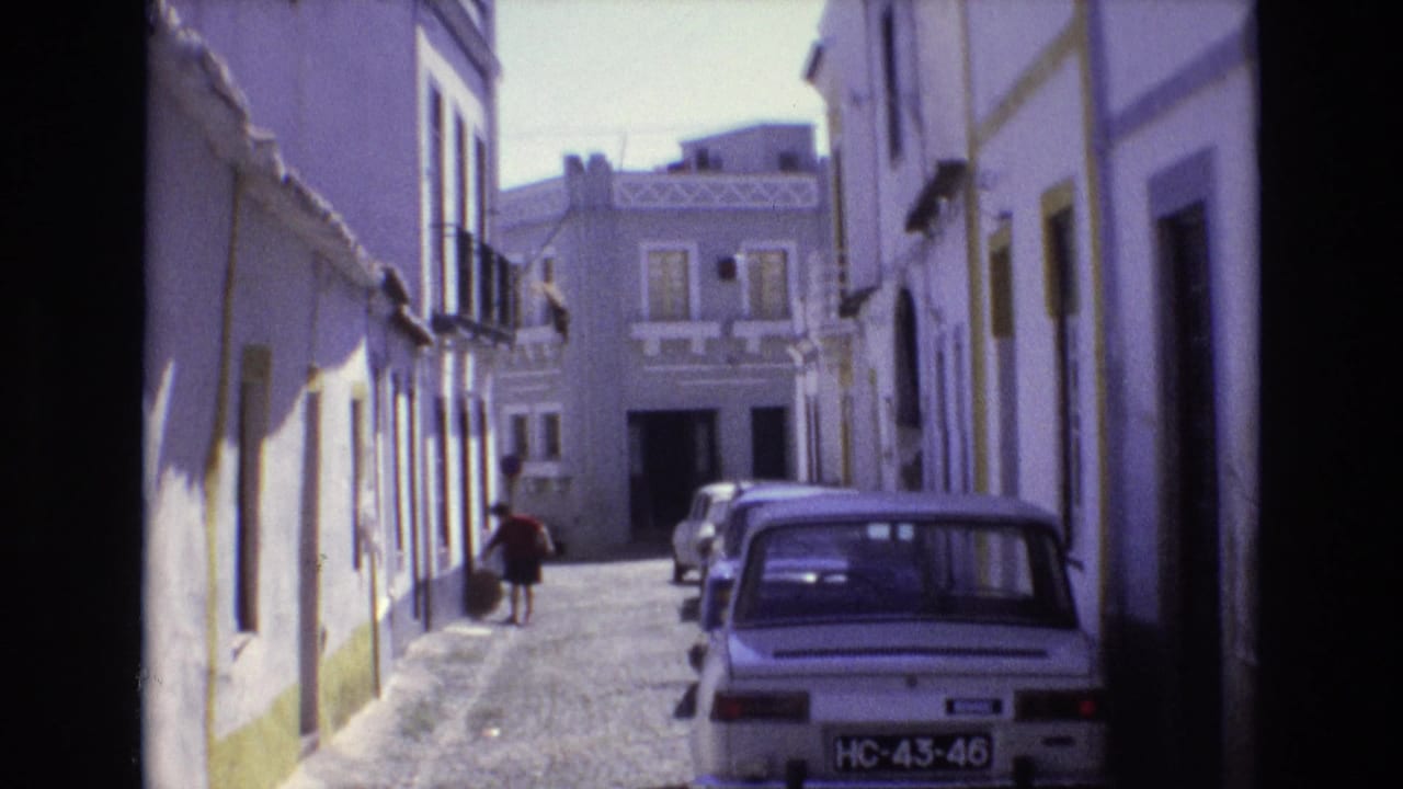 1972 Portugal 8mm frame showing a narrow cobblestone street lined with white buildings, yellow trim, balconies, parked cars, and a pedestrian walking away from the camera.