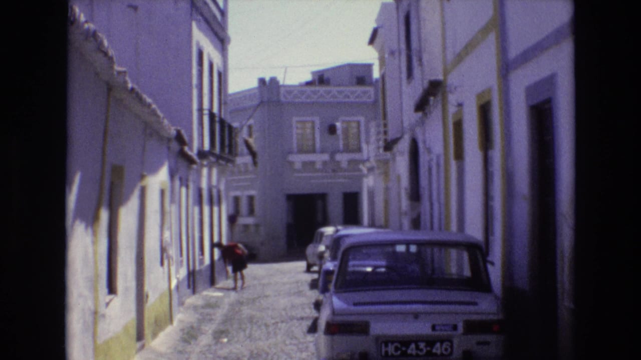 1972 Portugal 8mm frame looking down a narrow cobblestone lane with whitewashed facades, parked cars, balconies, and a person bending near a doorway on the left side.