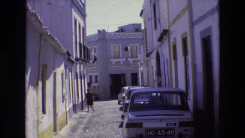 1972 Portugal 8mm frame showing a woman in dark clothing walking along a narrow cobblestone street bordered by white buildings with yellow trim and parked classic cars.