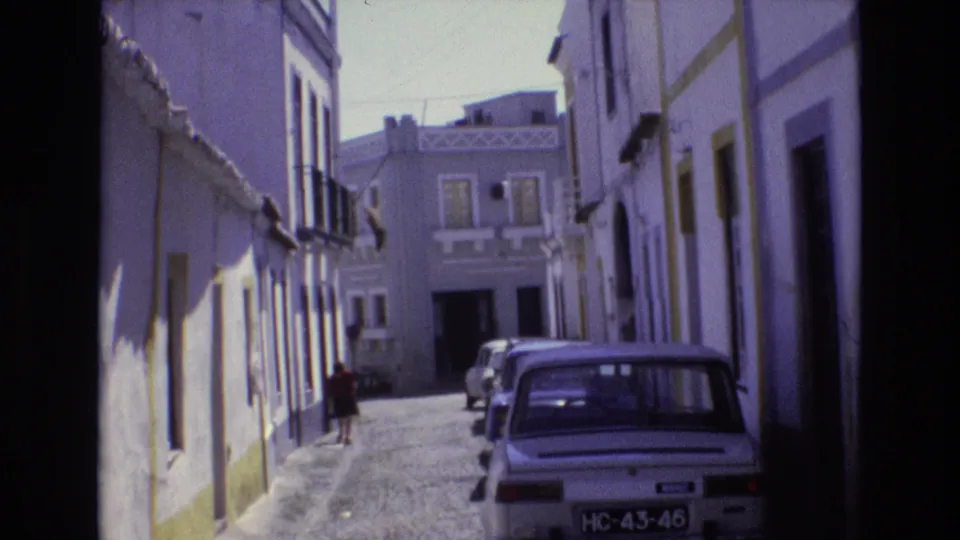 1972 Portugal 8mm frame of a quiet narrow cobblestone street with whitewashed buildings, yellow trim, parked cars, balconies, and a distant pedestrian near the left side.