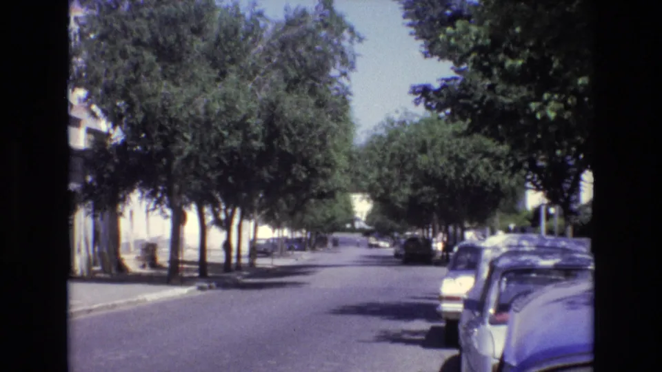1972 Portugal 8mm frame showing a quiet tree-lined street with parked classic cars, white building facades, and strong daylight on the road.