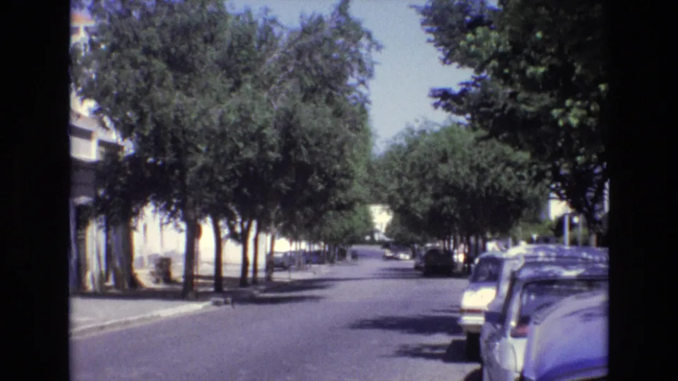 1972 Portugal 8mm frame showing a shaded residential street, leafy trees, parked cars at the curb, and white building exteriors in bright sun.