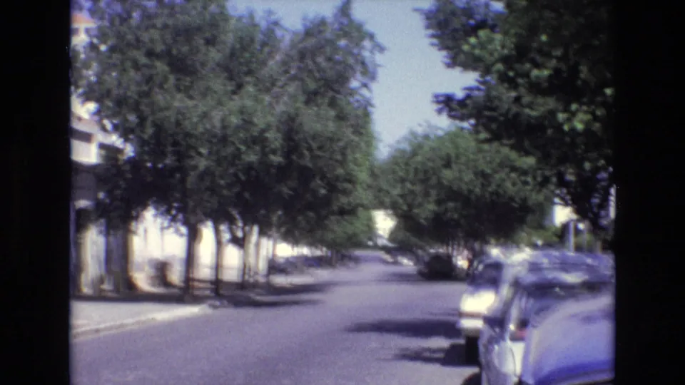 1972 Portugal 8mm frame with handheld blur across a quiet tree-lined street, parked cars, white facades, and dark scan borders.