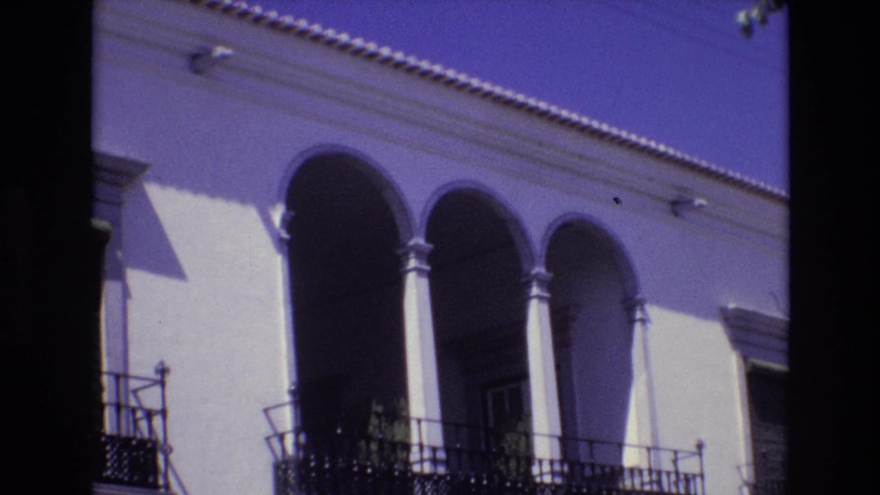1972 Portugal 8mm frame showing a white stucco building facade with rounded arches, slim columns, wrought-iron balcony railings, and deep shadows.