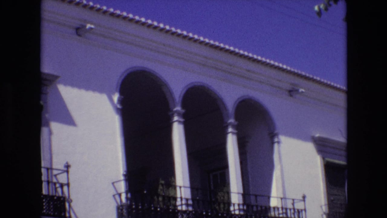 1972 Portugal home-movie frame of a white building with three arched openings, dark wrought-iron balcony railings, tiled roofline detail, and high-contrast sunlight.