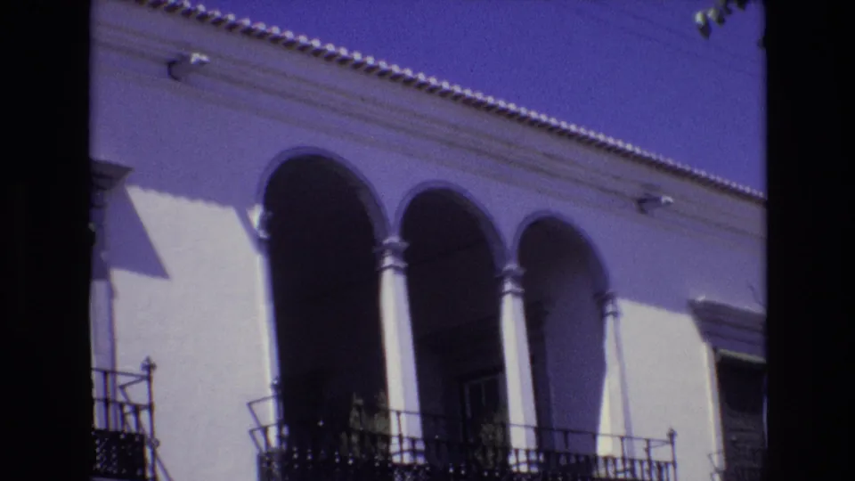 1972 Portugal home-movie frame of a white building with three arched openings, dark wrought-iron balcony railings, tiled roofline detail, and high-contrast sunlight.
